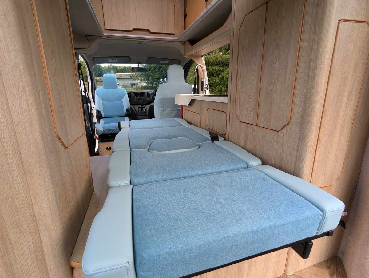 Micro campervan interior with light blue fold-out bed and wood cabinetry along walls