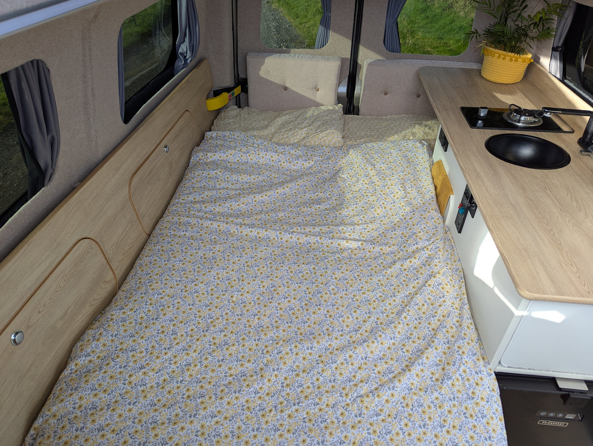 Camper van bed with patterned cover, side panels, and kitchen area on the right