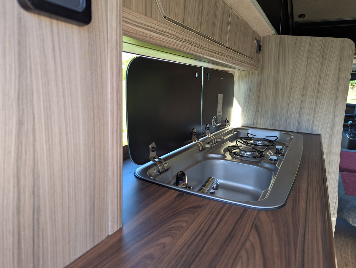 Campervan kitchen counter with stainless sink and two-burner stove in wood surround