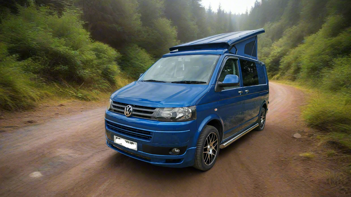 Blue VW camper van on a dirt road with pop-top roof folded down