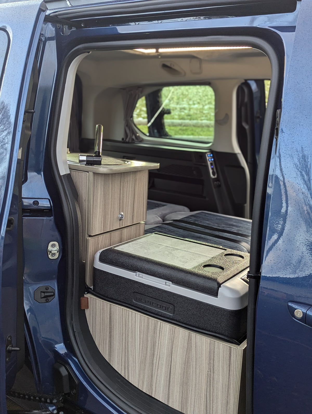 Campervan rear bed platform with grey cushions and wood cabinetry in open sliding area