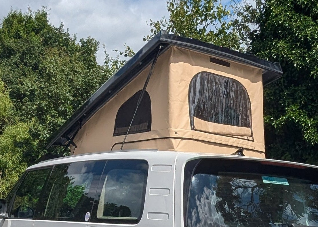 A Volkswagen ID. Buzz campervan van parked outdoors on a gravel driveway, featuring a beige rooftop tent and dark tree-like graphics on the rear side panel. Trees and shrubs surround the vehicle under a blue sky with white clouds.