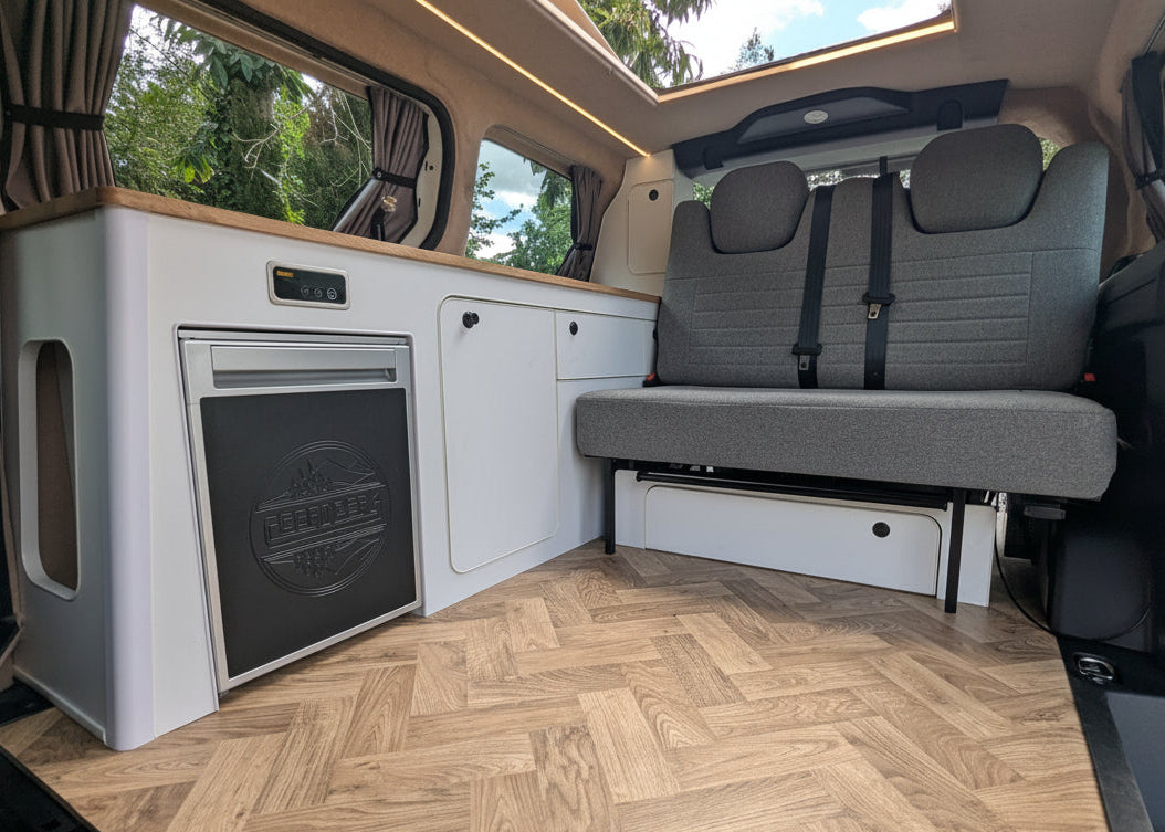 The interior of a Volkswagen ID. Buzz campervan showing a compact kitchenette with a white cabinet unit, a built-in black appliance, a gray fabric bench seat, and a light wood herringbone floor. A large window at the back shows trees and sky, and a sunroof is visible above.