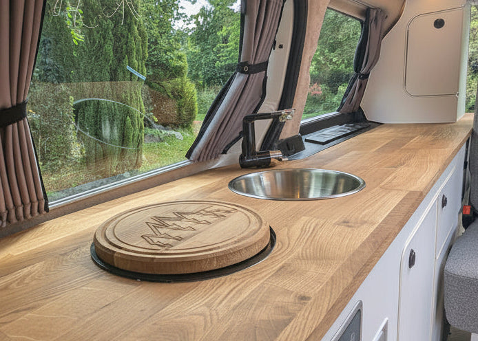 A compact kitchenette inside a Volkswagen ID. Buzz campervan, featuring a solid oak wood countertop with a built-in sink, a round wooden cutting board, brown curtains by a window, and a gray upholstered seat.