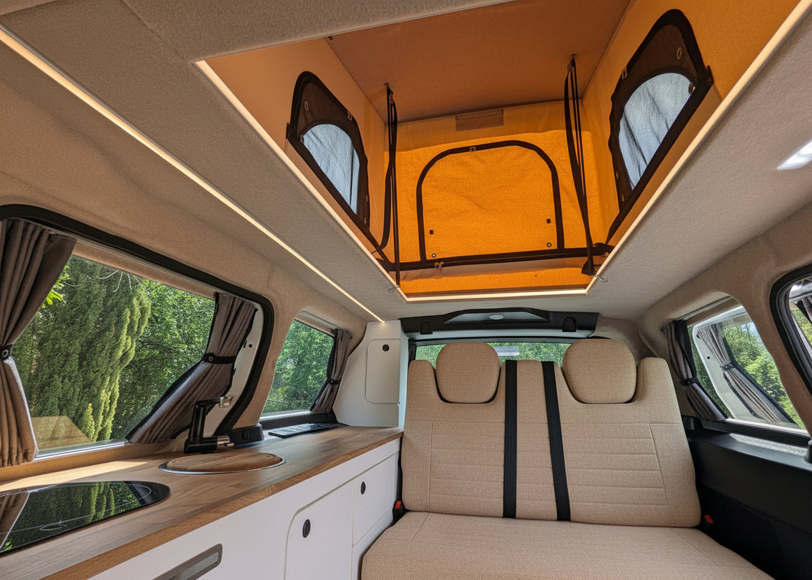 The interior of a Volkswagen ID. Buzz electric campervan showing a raised roof bed with an orange canopy top, gray fabric seats, a wood-look counter, and windows with curtains.
