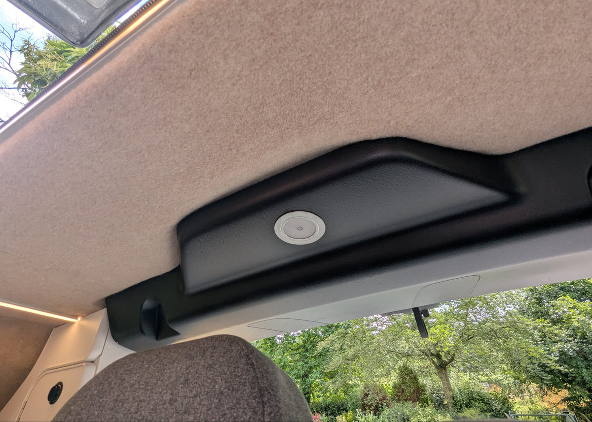 Close-up of the interior ceiling of a vw id buzz campervan, showing a beige carpeted overhead panel, a black plastic trim piece with a circular light or sensor, and a view of green trees through the window below.