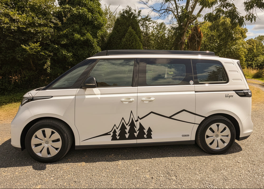 A white electric Volkswagen ID. Buzz campervan with a black mountain and pine tree graphic decal on the side, parked on a gravel path outdoors with trees and a blue sky in the background.