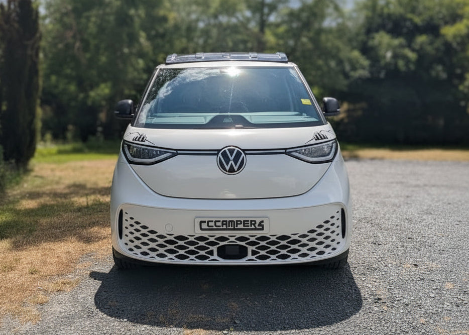 A white Volkswagen ID. Buzz electric campervan parked on a gravel path outdoors with trees and a cloudy sky in the background.