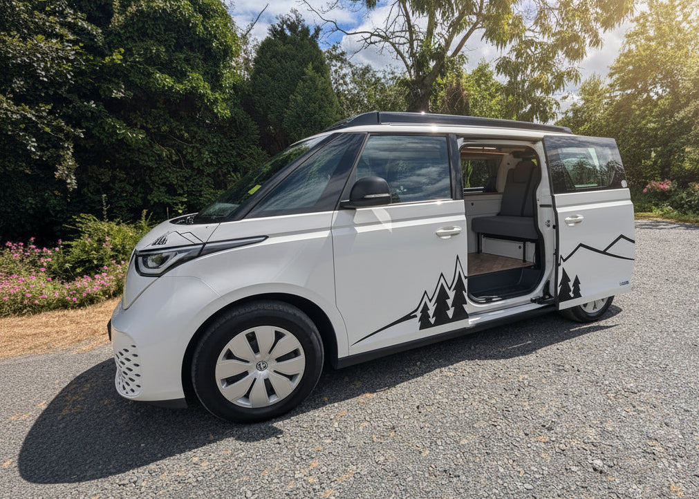 A white electric vw id buzz campervan with sliding side doors open, parked on a gravel surface outdoors with trees and a cloudy blue sky in the background. Black mountain and tree graphics decorate the side of the vehicle.