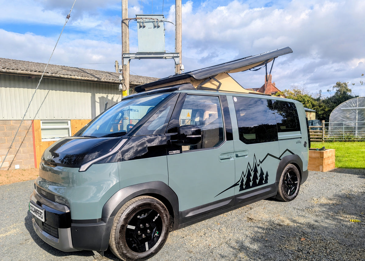 Front three-quarter exterior view of the grey Kia PV5 Witley campervan parked near a farm building with the pop-top roof open and modern styling