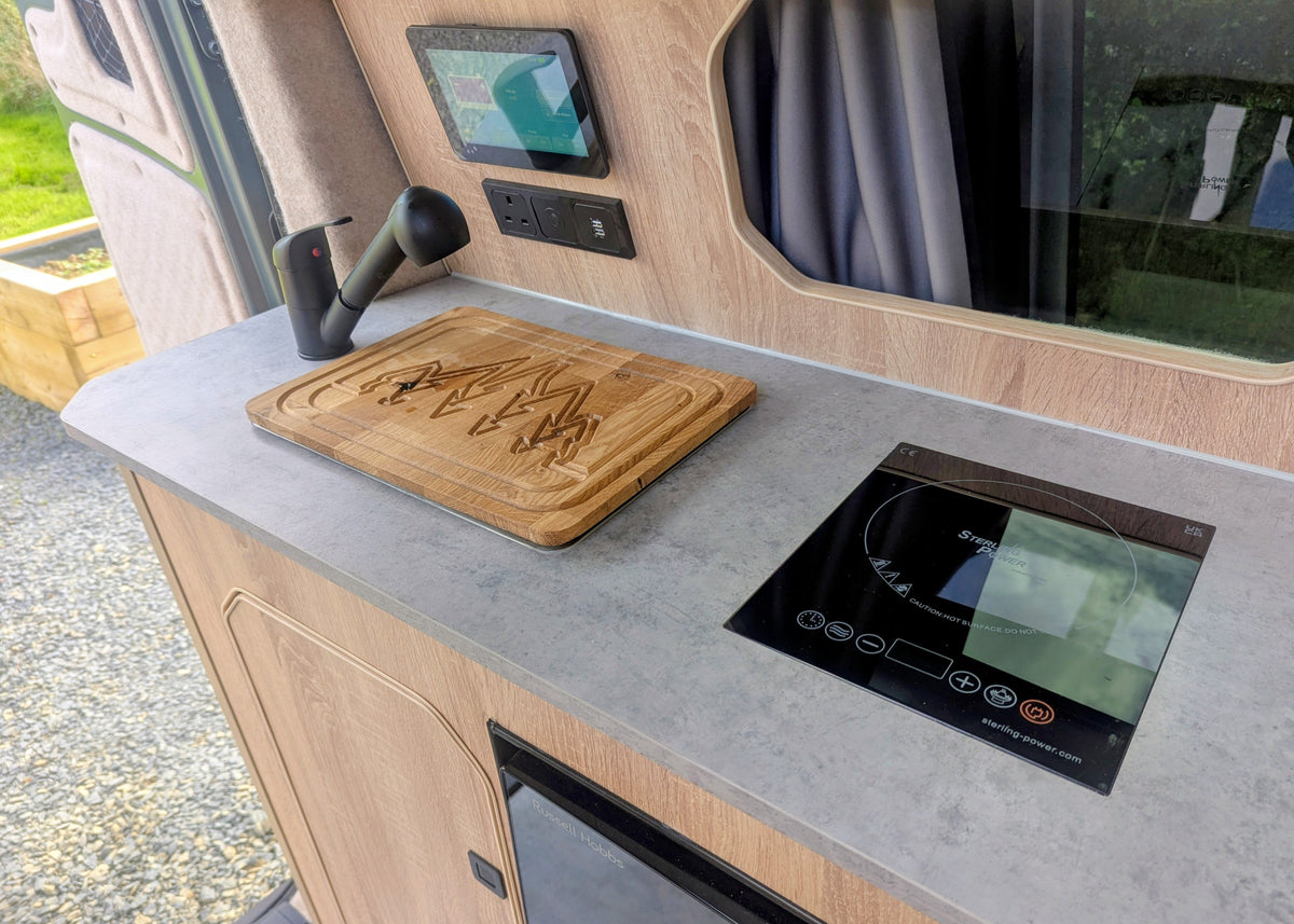 Angle shot of the Kia PV5 Witley kitchen worktop featuring a black sink tap, custom wooden chopping board sink cover, and portable induction cooktop.