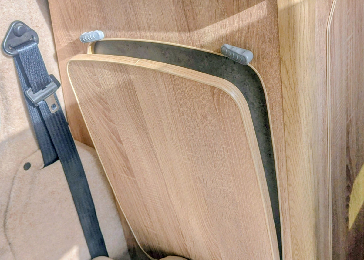 Interior view of the Kia PV5 Witley campervan showing the folded seat conversion or bed platform configuration next to the main kitchen cabinetry. WITH BUILT IN TABLE STORAGE
