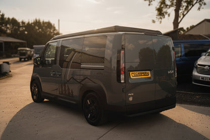 A Kia PV5 campervan parked outdoors on a concrete lot, shown from the rear three-quarter angle. The vehicle features tinted windows, black wheels, and a yellow 'CCCAMPERS' badge on the rear door.