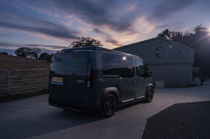 A dark gray Kia Cargo van parked on a concrete driveway in front of a green metal shed, with a field and trees in the background under a blue sky with clouds.