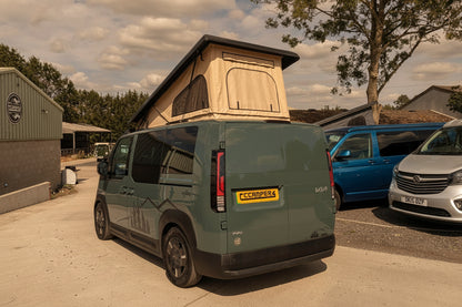 Olive Kia camper van with pop-top roof raised, rear three-quarter view