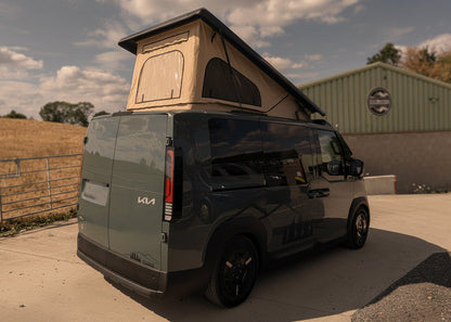 A Kia PV5 beige canvas pop top roof camper van with a rooftop tent raised above the roof, parked outdoors on a concrete surface near a green building under a blue sky with clouds.