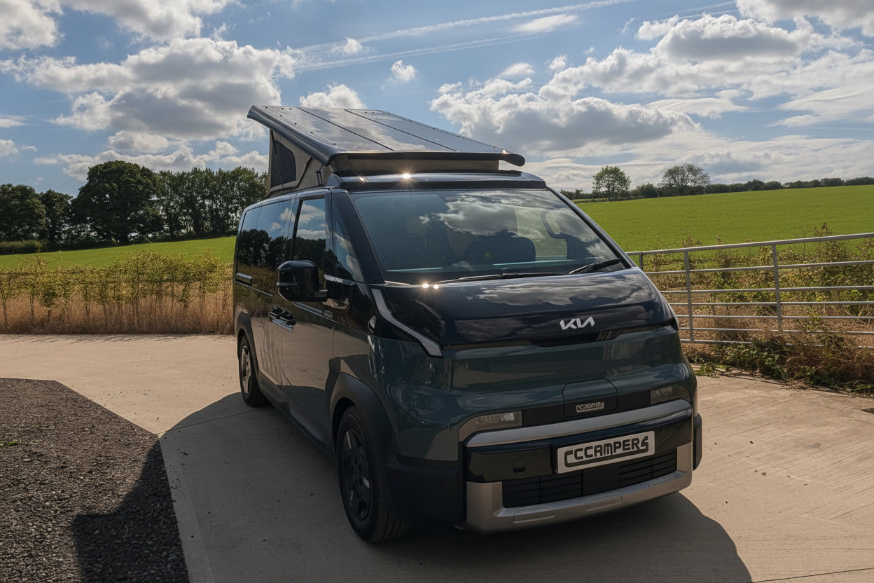 A Kia PV5 beige canvas pop top roof camper van with a raised roof and a mounted solar panel, parked on a concrete driveway outdoors under a blue sky with clouds.