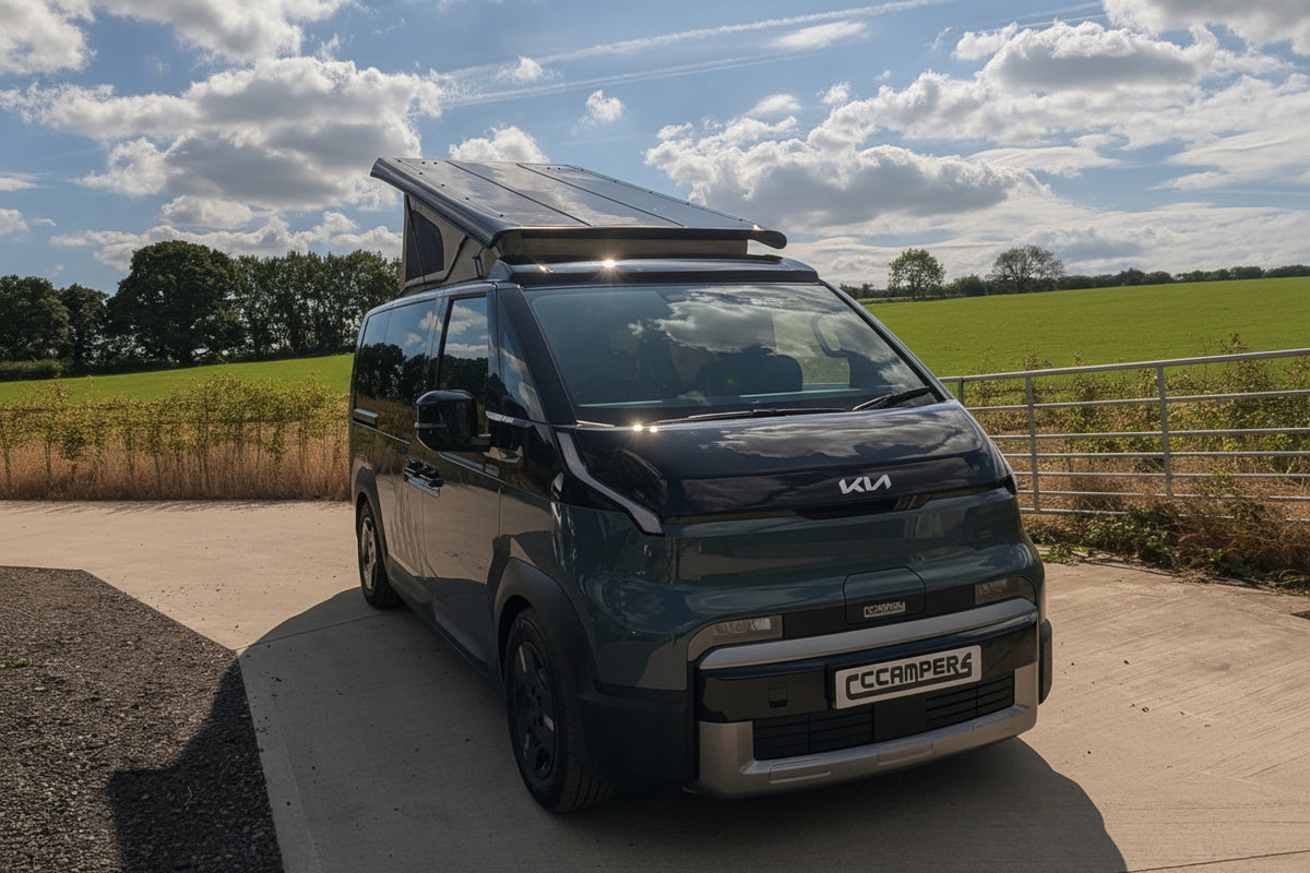A Kia PV5 beige canvas pop top roof camper van with a raised roof and a mounted solar panel, parked on a concrete driveway outdoors under a blue sky with clouds.