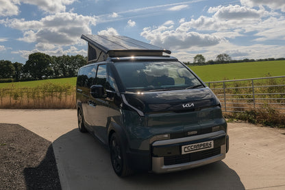 A Kia PV5 beige canvas pop top roof camper van with a raised roof and a mounted solar panel, parked on a concrete driveway outdoors under a blue sky with clouds.