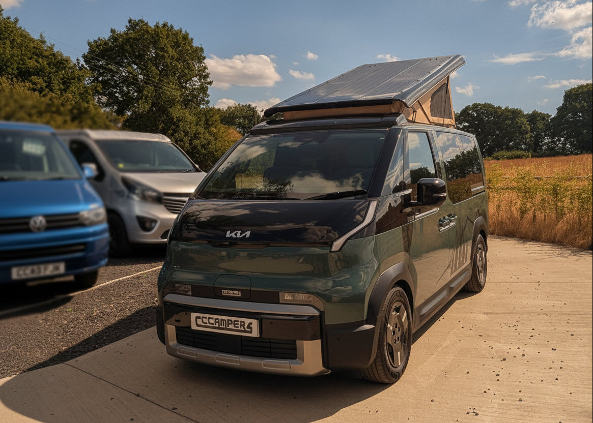 A dark green Kia PV5 camper van with a rooftop solar panel is parked on a concrete driveway outdoors. Other cars are visible in the background, along with trees, a field, and a blue sky with clouds.