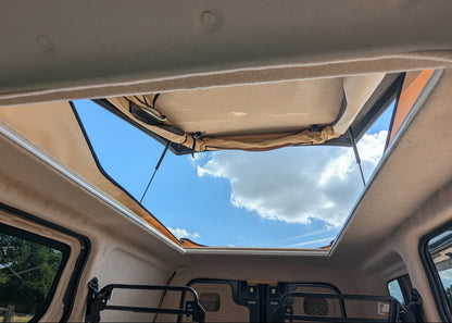 The interior rear area of a Kia PV5 with  a large open roof window looking out to the blue sky and clouds.