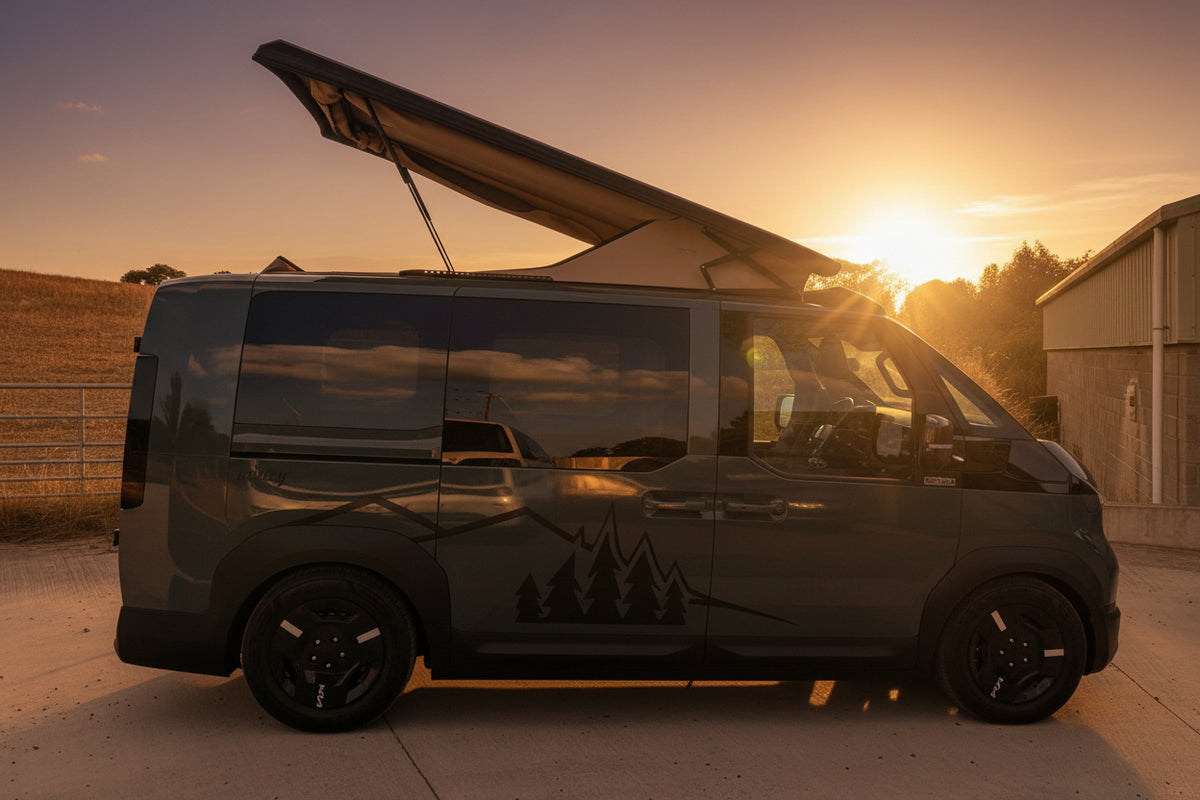 AKia PV5 beige canvas pop top roof  camper van with its roof pop-up tent extended, parked outdoors on a concrete surface under a blue sky with clouds. A building and dry grass are visible in the background, and part of a blue vehicle is in the bottom right corner.