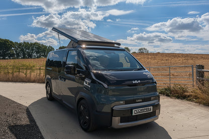 A dark gray Kia PV5 camper van with a solar panel on top, parked on a concrete driveway outdoors under a blue sky with clouds. A blue car is partially visible on the left, and a field with trees is in the background.