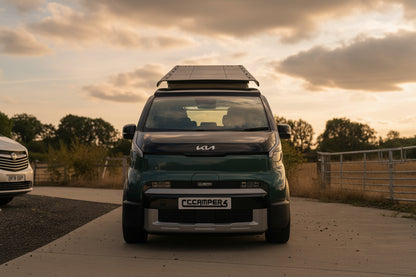 A dark green Kia PV5 campervan parked on an outdoor concrete driveway under a blue sky with clouds, with other cars and a field visible in the background.