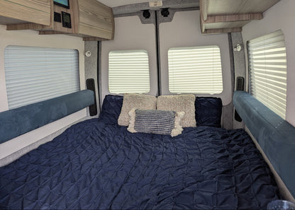 A cozy bedroom setup inside a camper or RV, featuring a dark navy blue quilted bedspread, matching pillows, textured cream and striped accent pillows, and blue upholstered side benches under windows with white blinds.