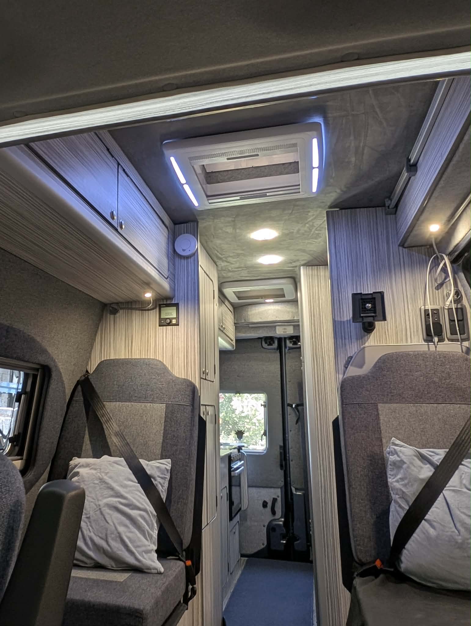 The interior of a motorhome or camper van, showing two gray upholstered passenger seats with seatbelts, light-colored wood-look cabinets on both sides, a blue-gray carpeted aisle, and a ceiling-mounted air conditioner with integrated LED lighting.