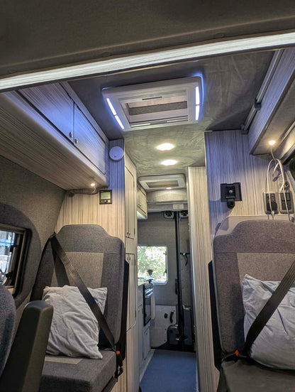 The interior of a motorhome or camper van, showing two gray upholstered passenger seats with seatbelts, light-colored wood-look cabinets on both sides, a blue-gray carpeted aisle, and a ceiling-mounted air conditioner with integrated LED lighting.