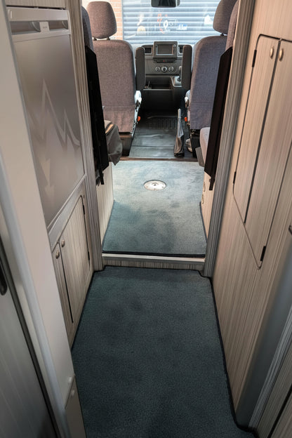 The interior hallway of a recreational vehicle or motorhome, showing gray carpeted floors, light wood-paneled walls, and gray upholstered seats leading toward a front cabin area.