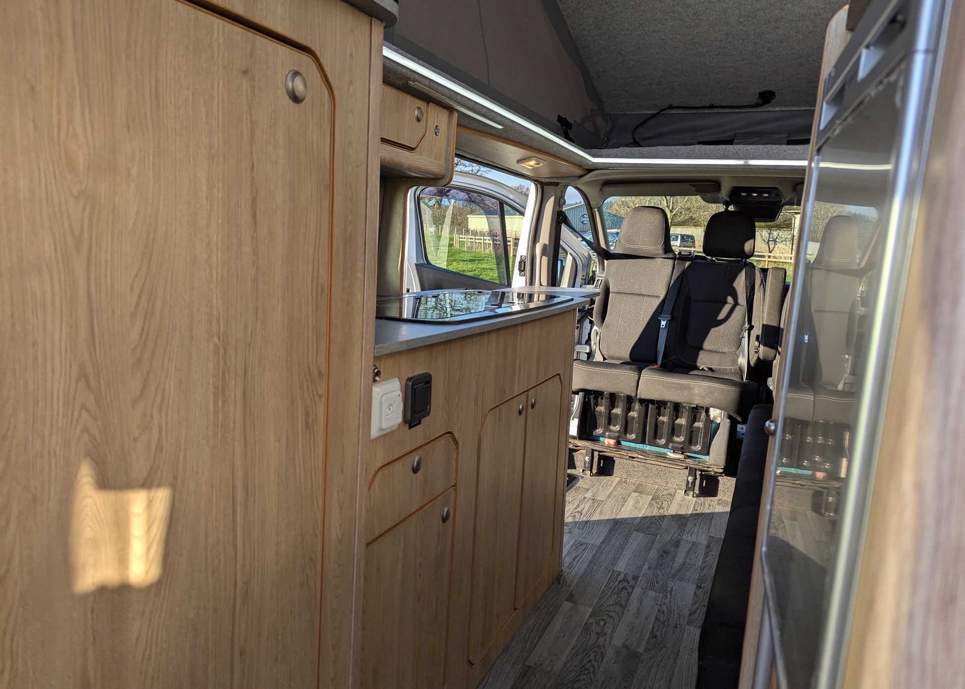 The interior side of a camper van or motorhome showing light wood cabinetry, a countertop, a sink area, and the rear seating area with a black seat in the background.