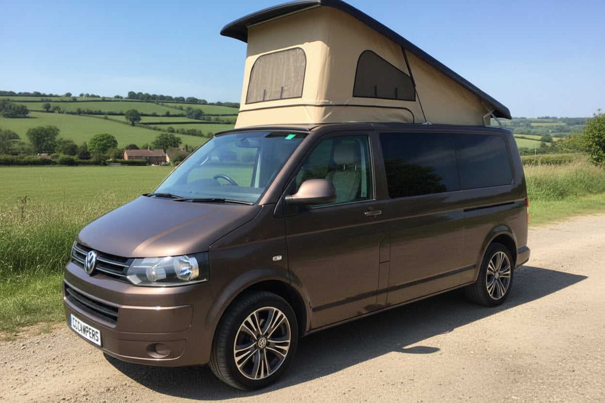A dark brown Volkswagen camper van with a beige pop-up caravan roof, parked outdoors on a gravel driveway with trees and a house in the background.