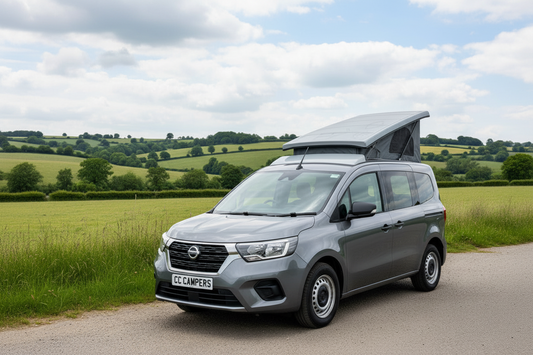 A gray Nissan-Townstar with a rooftop tent popped open, parked on a country road with green fields and hills under a cloudy sky.