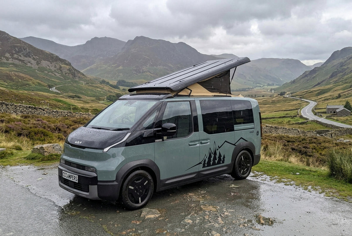 Premium CCCAMPERS Kia PV5 electric campervan conversion built for UK road trips, parked on a scenic mountain pass road in the Scottish Highlands. The custom eco-friendly camper features a distinctive black and sage green two-tone paint finish, custom geometric mountain decals, gloss black alloy wheels, and an open pop-top roof tent with a breathable tan canvas. This cutting-edge, purpose-built electric van conversion highlights the best of British craftsmanship, showcasing a practical, off-grid adventure-re