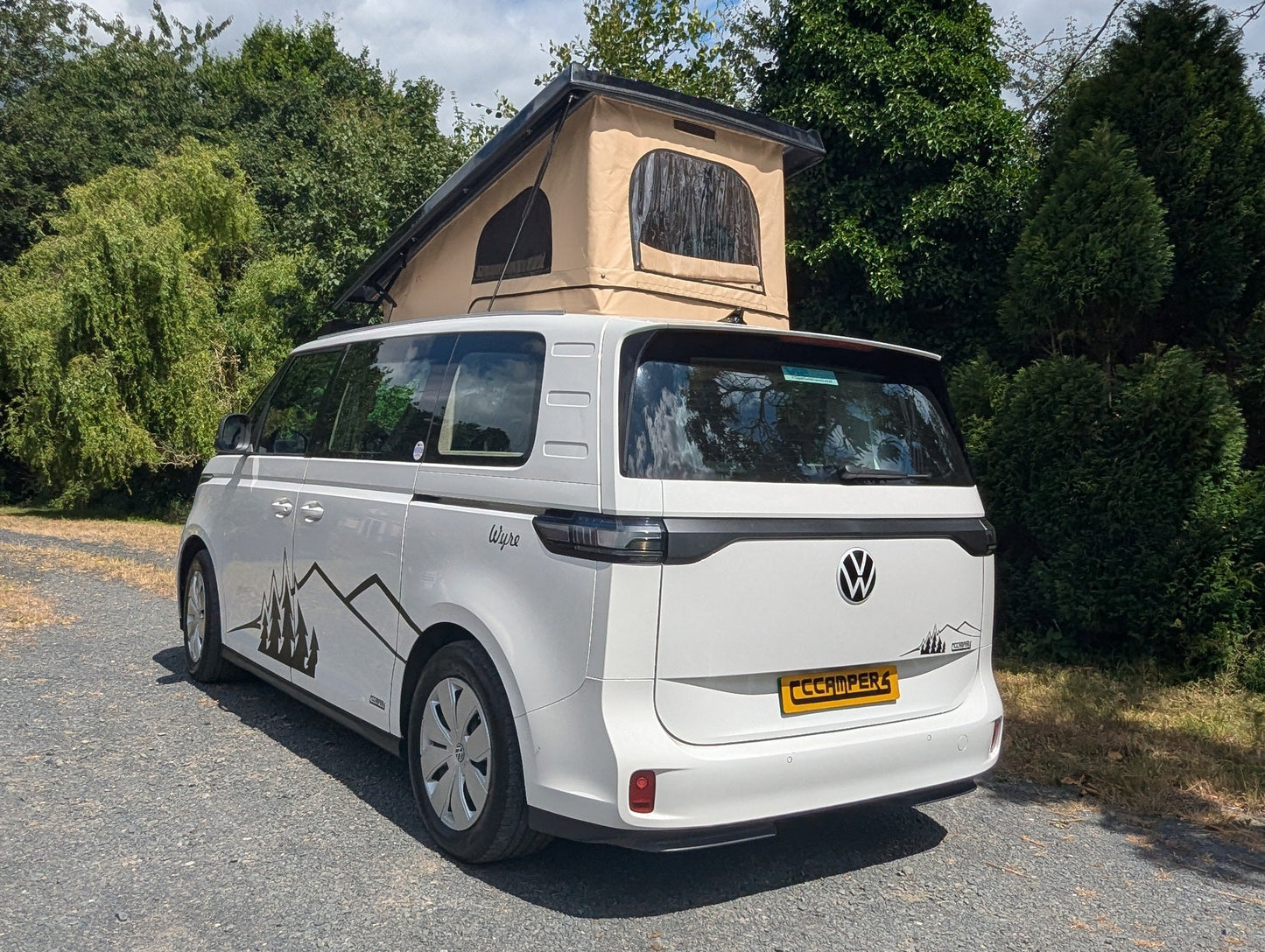 A Volkswagen ID. Buzz campervan van parked outdoors on a gravel driveway, featuring a beige rooftop tent and dark tree-like graphics on the rear side panel. Trees and shrubs surround the vehicle under a blue sky with white clouds.