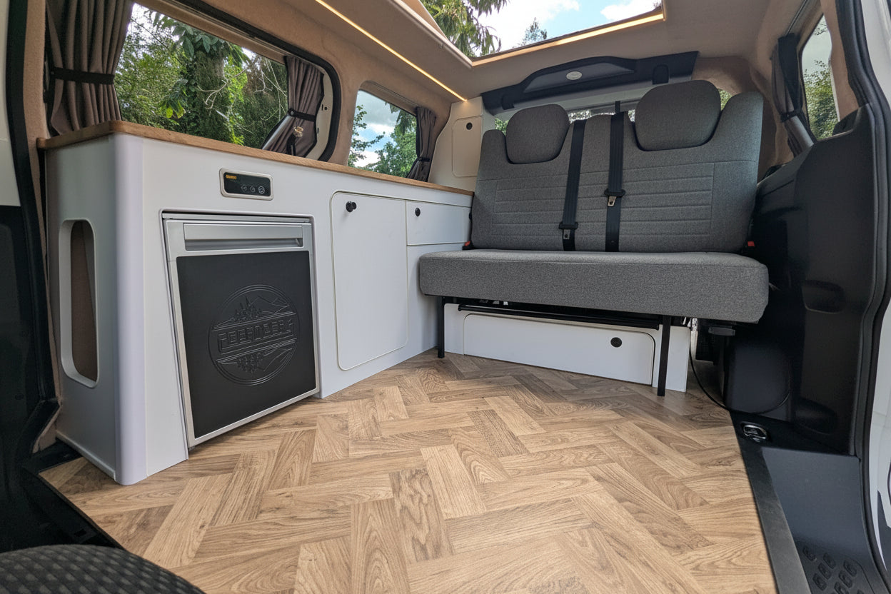 The interior of a Volkswagen ID. Buzz campervan showing a compact kitchenette with a white cabinet unit, a built-in black appliance, a gray fabric bench seat, and a light wood herringbone floor. A large window at the back shows trees and sky, and a sunroof is visible above.