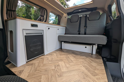 The interior of a Volkswagen ID. Buzz campervan showing a compact kitchenette with a white cabinet unit, a built-in black appliance, a gray fabric bench seat, and a light wood herringbone floor. A large window at the back shows trees and sky, and a sunroof is visible above.