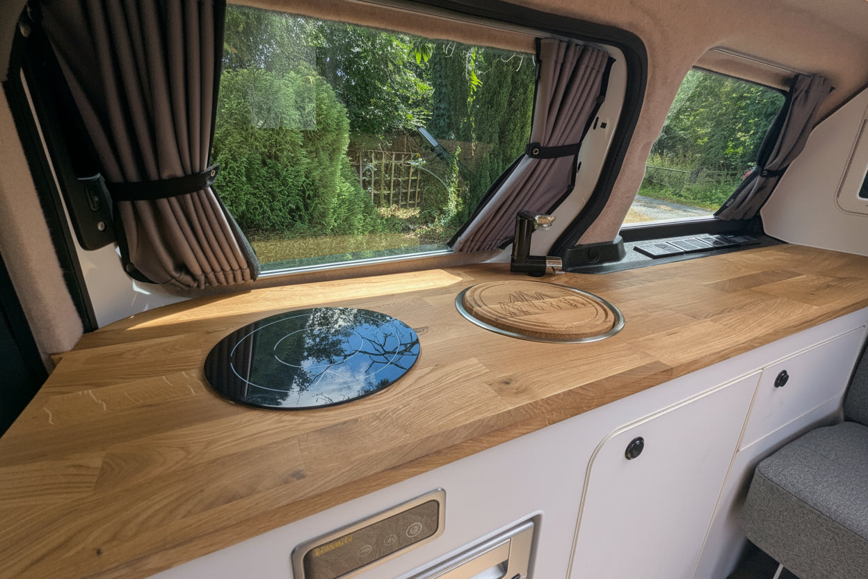 A close-up view of a wooden kitchenette counter inside a Volkswagen ID. Buzz campervan, featuring a light oak wood countertop with an integrated black circular cooktop and a round wooden tray, set against white cabinets and large windows with curtains.