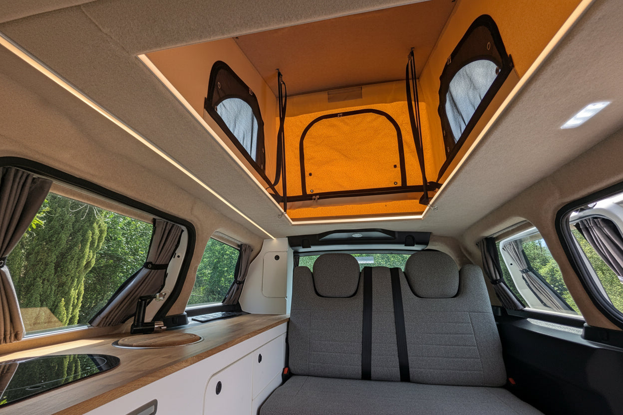The interior of a Volkswagen ID. Buzz electric campervan showing a raised roof bed with an orange canopy top, gray fabric seats, a wood-look counter, and windows with curtains.