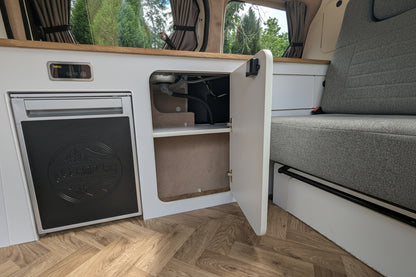 A vw id buzz camper kitchen interior with a white refrigerator, open storage cabinet, wood flooring, and a gray upholstered bed seat beside it.