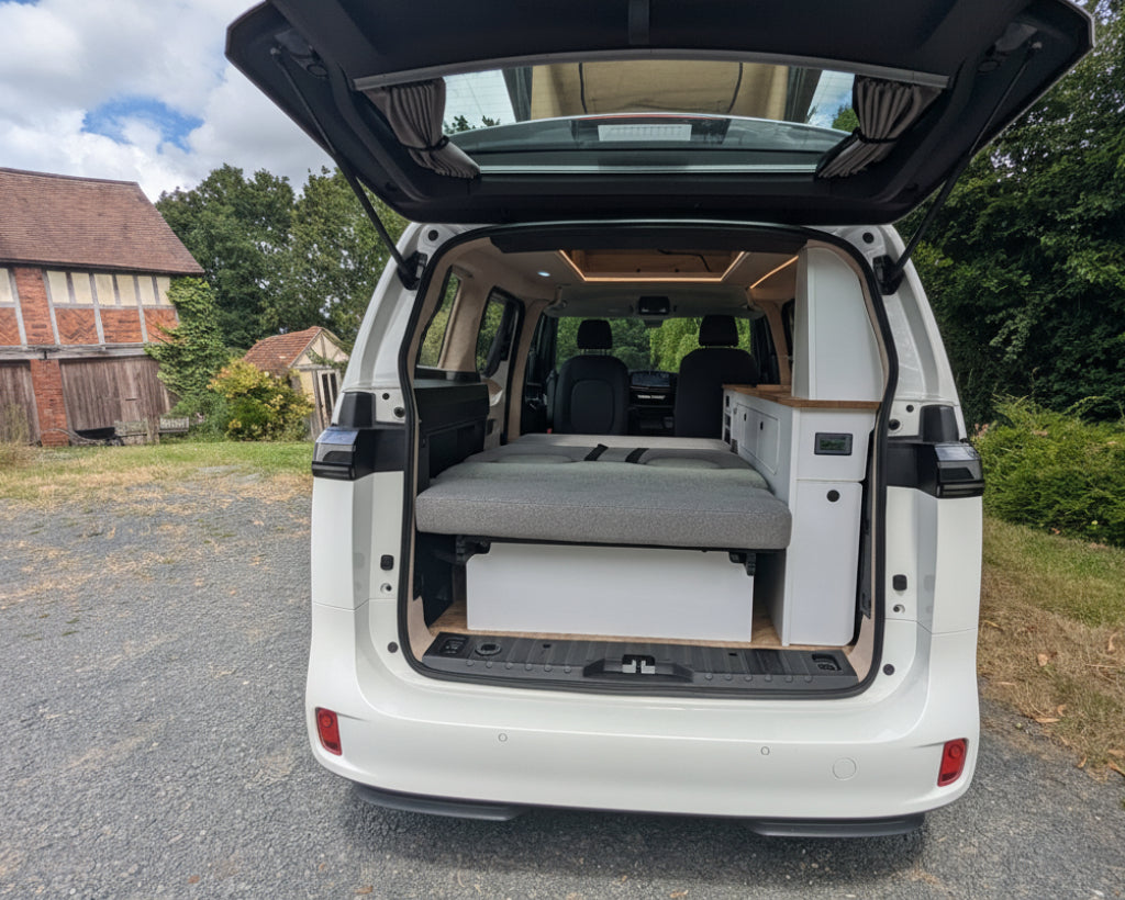 A white vw id buzz with its rear hatch open, showing a custom camper setup inside with a gray bed platform and storage compartments. The vehicle is parked outdoors on a gravel surface with trees, grass, and a brick building in the background.