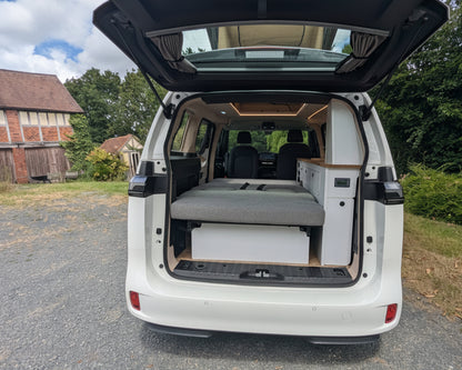 A white vw id buzz with its rear hatch open, showing a custom camper setup inside with a gray bed platform and storage compartments. The vehicle is parked outdoors on a gravel surface with trees, grass, and a brick building in the background.