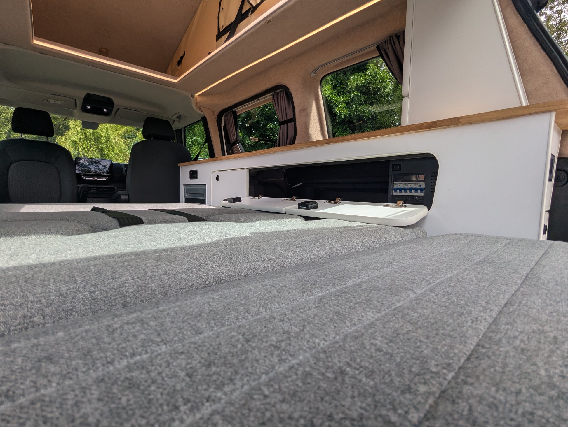 A low-angle view inside a  vw id buzz camper van showing bed down, with easy access white cabinetry, wood trim, windows, and green foliage visible outside.