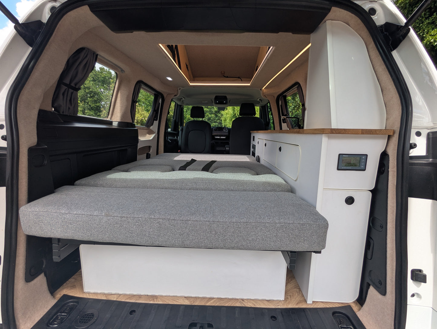 The rear cargo area of a  vw id buzz converted into a camper, showing a raised gray fabric bed frame, white storage cabinets, wood-look trim, and the vehicle's interior.