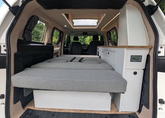 The rear cargo area of a  vw id buzz converted into a camper, showing a raised gray fabric bed frame, white storage cabinets, wood-look trim, and the vehicle's interior.