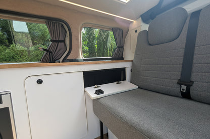 Gray upholstered travellin 3/4 seat with integrated headrest and seatbelt, shown inside a vw id buzz with windows, curtains, and a white storage cabinet beside it.