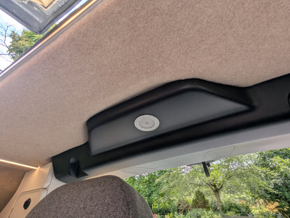 Close-up of the interior ceiling of a  vw id buzz campervan, showing a beige carpeted overhead panel, a black plastic trim piece with a circular light or sensor, and a view of green trees through the window below.