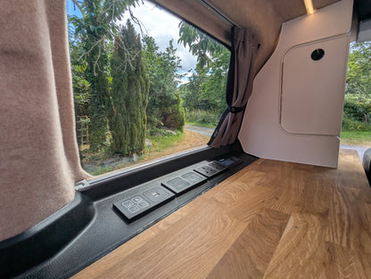 Close-up of a vw id buzz camper van interior with a wood-look countertop, window controls, curtains, and an outdoor view of green trees and a path.